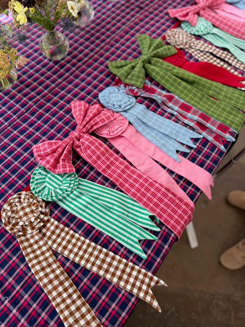 A collection of colorful fabric bow and ribbon accessories in various plaid and striped patterns, arranged diagonally on a red and blue plaid tablecloth.