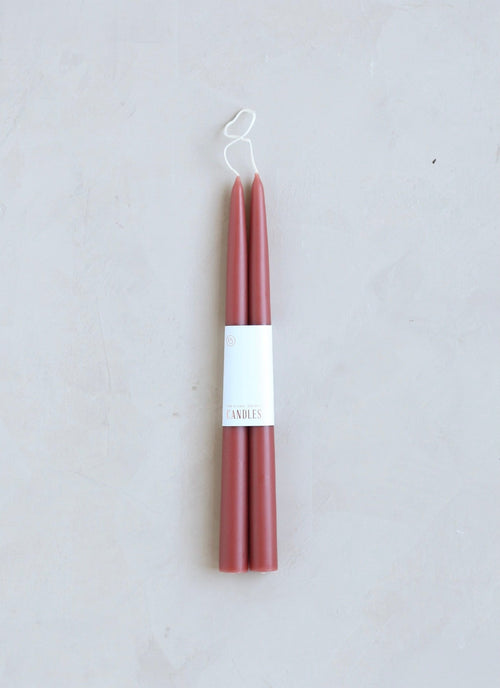 Two red taper candles with a white label in the center, placed on a neutral background.