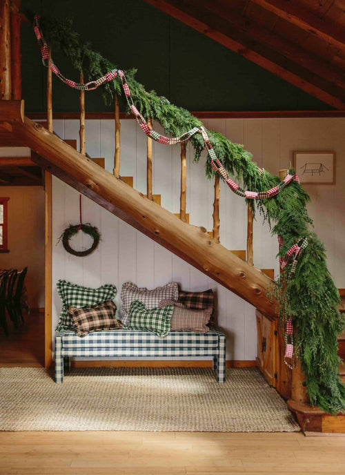 A festive garland with red, green, and multicolor patterns draped over a staircase, with a bench and greenery in the background.