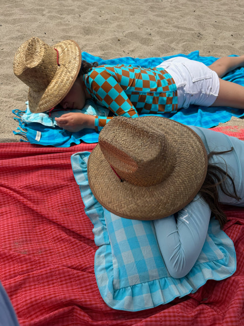 Two people lying on beach towels with hats on, one person wearing a checkered shirt and white shorts.