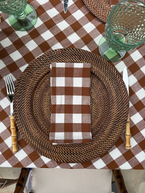 A styled dining table setting featuring a brown and white gingham checkered tablecloth, a brown woven charger plate, a matching folded napkin, bamboo-handled cutlery, and green textured glassware.