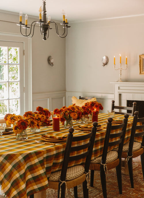 A Gingham Oak tablecloth laid out on a dining table, with a decorative hand-stitched center seam, surrounded by a set of chairs in a cozy room with autumn decor.