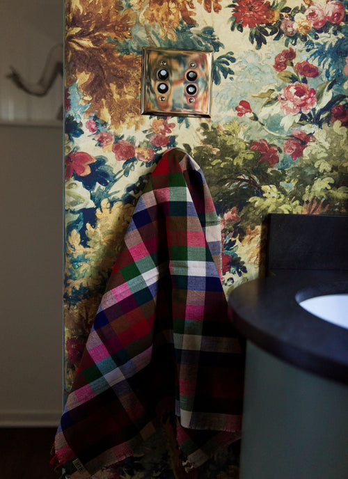 A multicolor plaid bath towel hangs from a hook on a wall covered in vintage floral wallpaper. A dark countertop and a portion of a sink are visible on the right side.