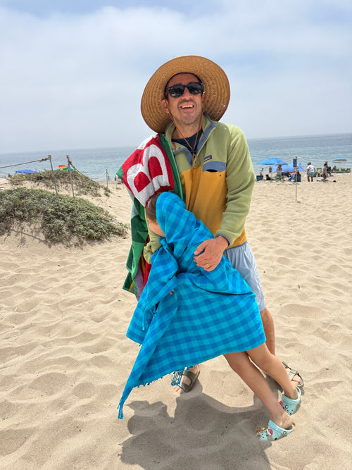 Person on a beach holding a blue beach towel with a colorful pattern, wearing a straw hat and sunglasses.