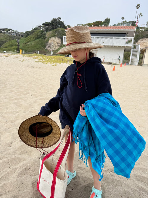 Person on a beach holding a hat, towel, and tote bag with a building and greenery in the background.
