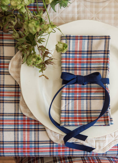 A folded plaid napkin with a navy blue ribbon bow, arranged on a white plate over a matching plaid tablecloth and beige charger plate, with dried green flowers in the corner.
