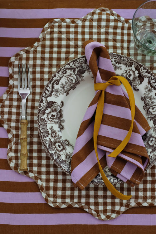 A styled place setting featuring a brown and white gingham placemat with a scalloped edge, a brown and white floral plate, a pink and brown striped napkin tied with a yellow ribbon, and a pink and brown striped tablecloth.