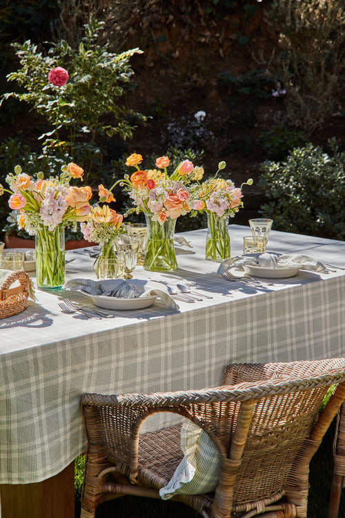 An outdoor dining table setting featuring a green plaid tablecloth, neatly laid out with plates and accompanied by floral centerpieces in glass vases.