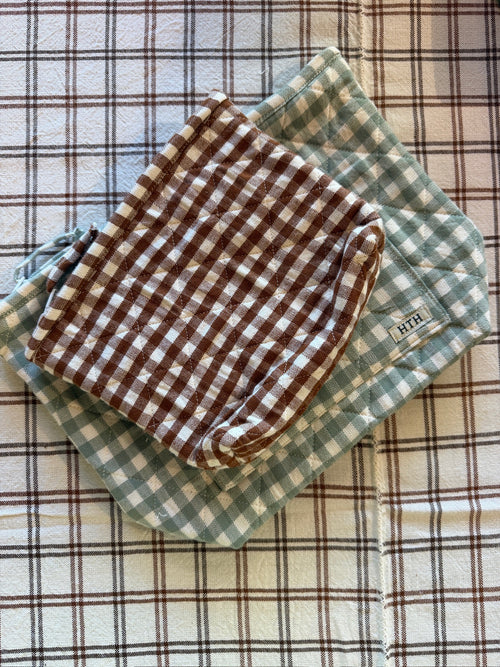 Two folded square cloth pouches with gingham checks, one brown and white and one sage green and white, stacked on a beige plaid fabric background. A small fabric tag is visible on the green cloth.