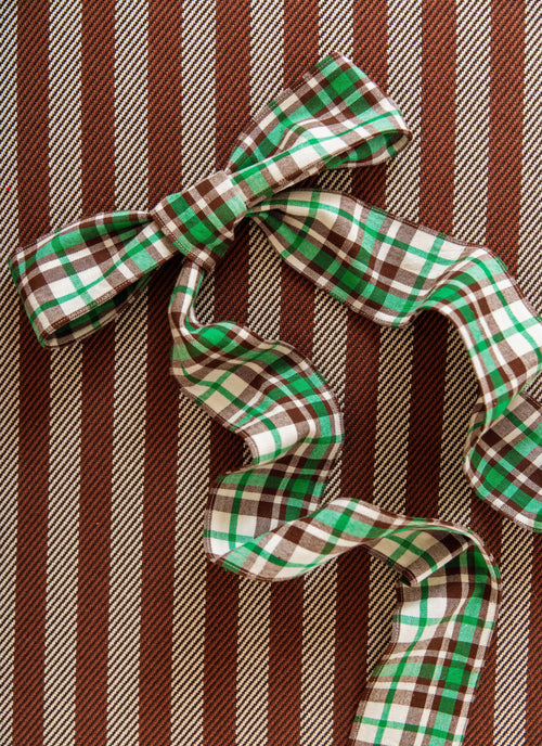 A green, brown, and white plaid ribbon tied into a bow, laid flat on a brown and beige vertically striped fabric background.