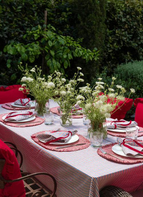 Dining table set with red and white tablecloth, floral centerpieces, and red chairs outdoors.