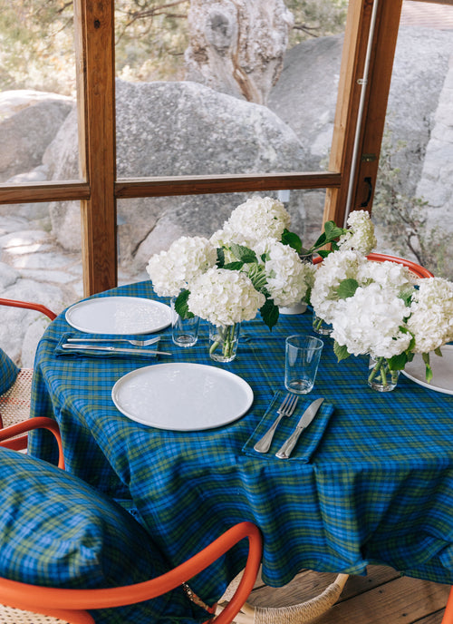 Table setting with blue plaid tablecloth, white plates, silverware, and floral centerpieces in a natural setting.