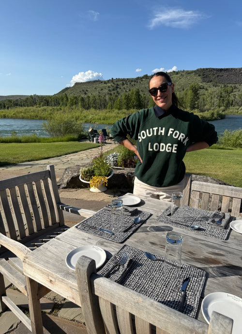 Person wearing a 'South Fork Lodge' shirt standing by an outdoor dining table with a scenic background.