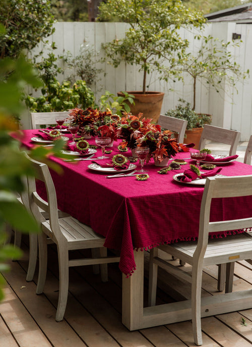 Outdoor dining table set with a red tablecloth, floral centerpieces, and white chairs.