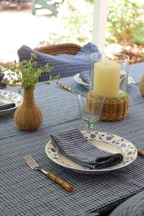 A styled outdoor dining table setting with a blue and white gingham tablecloth, matching napkins on floral plates, a small wicker vase with greenery, and a glass candle holder.