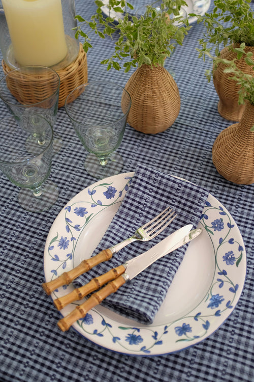 A styled tabletop setting featuring a blue and white gingham tablecloth, a white plate with a blue floral border, a blue and white checkered napkin, and a silver knife and fork with bamboo handles. Decorative wicker baskets, glass candle holders, and small potted green plants are arranged around the setting.