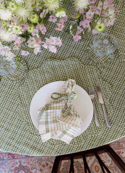 Table setting with a white plate, checkered napkin, and cutlery on a green checkered tablecloth.