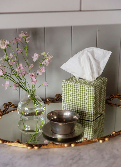 Green checkered tissue box with white tissues on a mirror surface with flowers and a bowl.