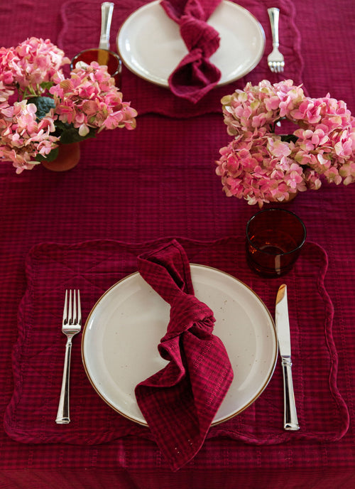 Dining table setting with purple napkins, pink flowers, and silverware on a red tablecloth.