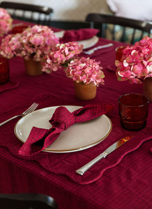 Dining table set with purple red napkins, plates, and flowers on a red tablecloth.