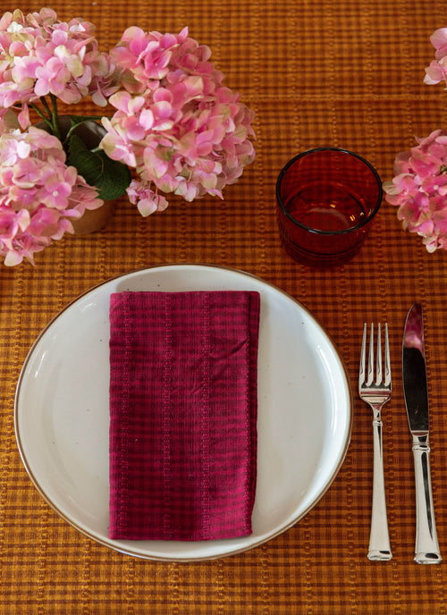 Table setting with a white plate, purple checkered napkin, fork, knife, and pink flowers on an orange placemat.