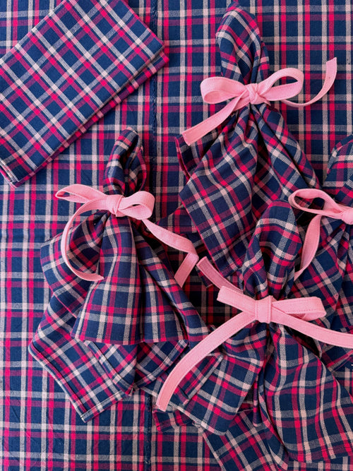 A close-up flat lay of matching plaid fabric accessories, including a folded napkin, a folded napkin tied with a pink ribbon, and a folded cloth tied with a pink ribbon, all on top of a larger matching plaid fabric background.